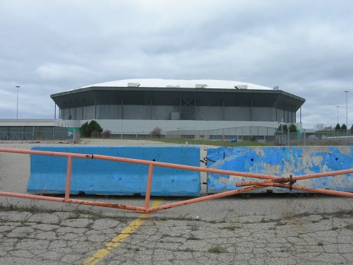 An abandoned stadium viewed from a distance, with a barrier in the foreground and a cloudy sky above.