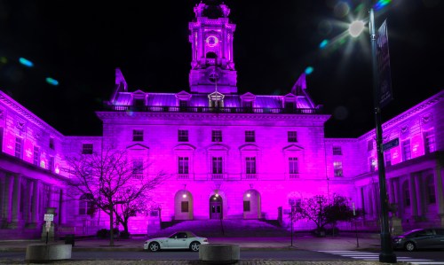 Historic building illuminated in vibrant purple light at night, showcasing its architectural details and surrounding trees.