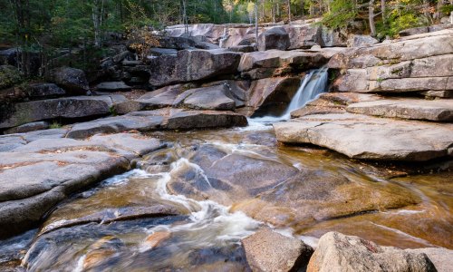 A serene landscape featuring a flowing stream over large rocks, surrounded by trees in a tranquil forest setting.
