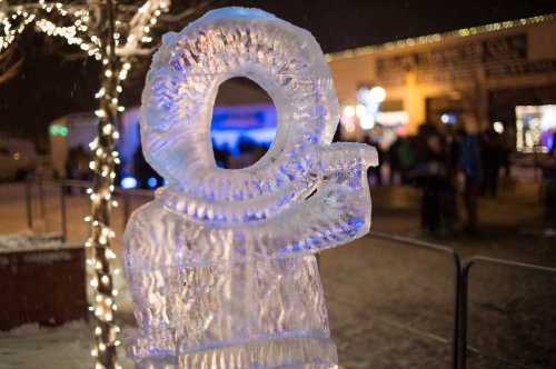 A large ice sculpture with a circular design, illuminated by blue lights, set in a snowy outdoor festival scene.