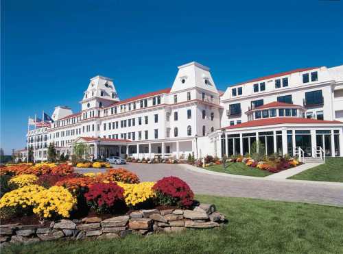 A large white hotel with red roofs, surrounded by colorful flower beds and a clear blue sky.