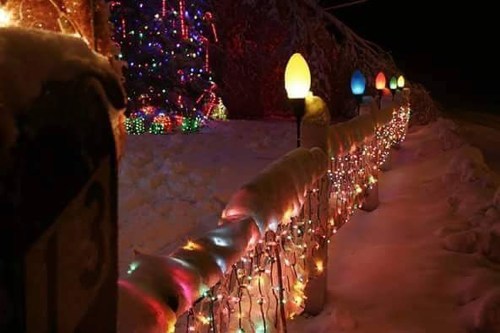 A snowy pathway lined with colorful lights and a decorated tree, creating a festive winter scene at night.