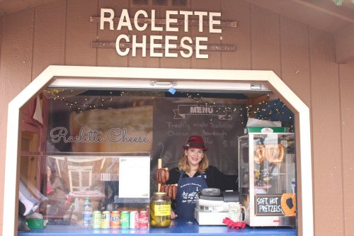 A smiling vendor stands behind a raclette cheese stall, with a menu and various food items displayed.