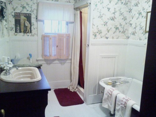 A vintage bathroom with floral wallpaper, a bathtub, sink, and a shower curtain, featuring a window with wooden shutters.
