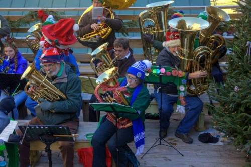 A festive group of musicians playing tubas and euphoniums, wearing holiday hats and surrounded by Christmas decorations.