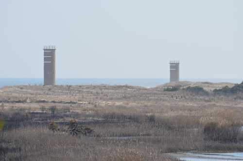 Two tall, cylindrical structures stand on a grassy landscape near the ocean, with a clear sky in the background.