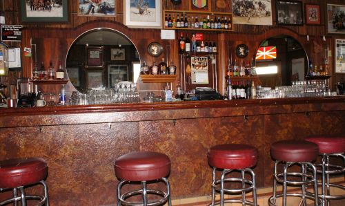 A wooden bar with red stools, mirrors, and shelves lined with various bottles and glasses, decorated with photos.