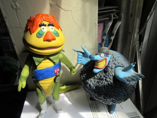 Two colorful, cartoonish characters stand side by side, one with orange hair and a green outfit, the other round and blue.