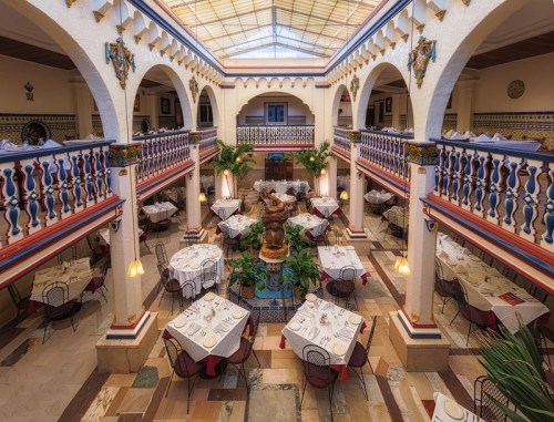 A spacious restaurant with elegant tables, arches, and a central fountain, surrounded by greenery and decorative tiles.