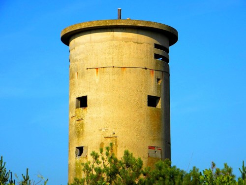 A weathered concrete tower with a circular top, surrounded by greenery against a clear blue sky.