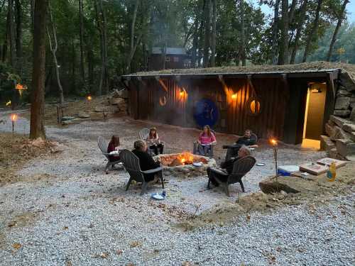 A group of people sitting around a fire pit in a wooded area, with a rustic building and torches in the background.