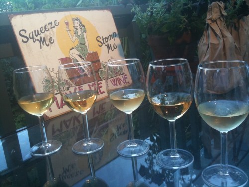 Five glasses of white wine on a table, with a vintage sign in the background that reads "Squeeze Me, Stomp Me, Make Me Wine."