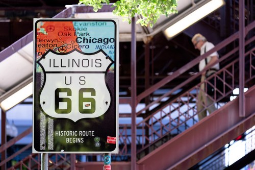 Sign for Route 66 in Illinois, indicating the historic route begins, with a person walking in the background.