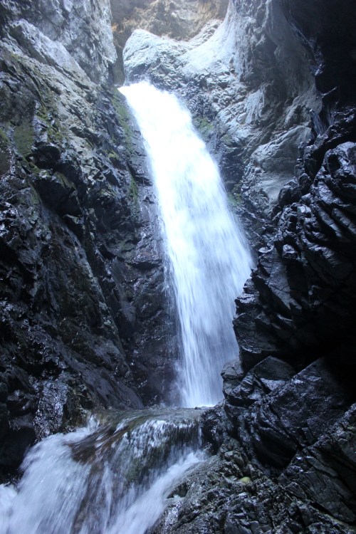 A cascading waterfall flows down rocky cliffs, surrounded by dark, textured stone and lush greenery.