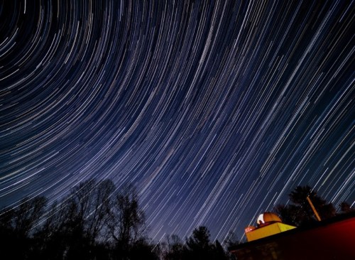Star trails swirl in the night sky above a silhouette of trees and a building, creating a dynamic celestial scene.
