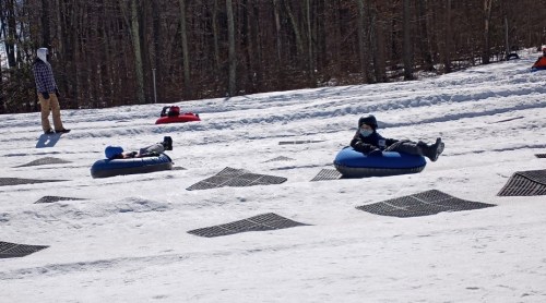 Two children in snow tubes slide down a snowy hill, while an adult stands nearby in winter attire.
