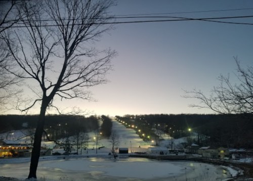 A serene winter landscape at dusk, featuring a snowy hill lined with lights and bare trees in the foreground.