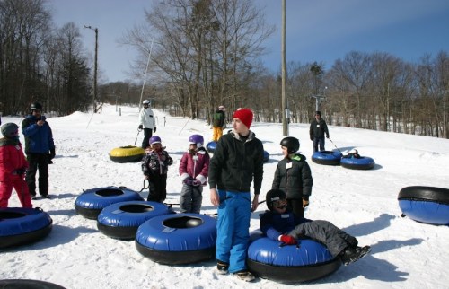 Children and adults enjoy tubing on a snowy hill, with ski lifts and trees in the background.