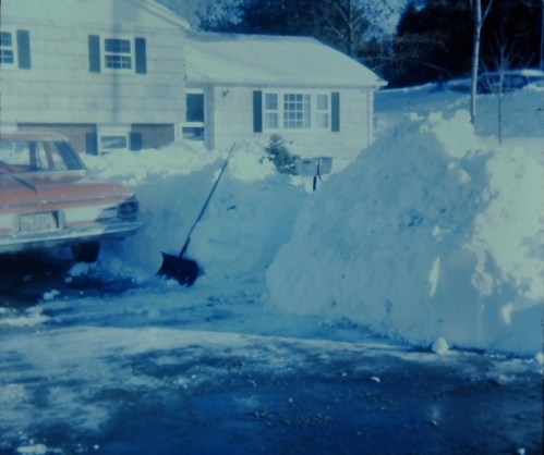 A snowy scene with a car parked beside large snow piles and a person shoveling snow near a house.