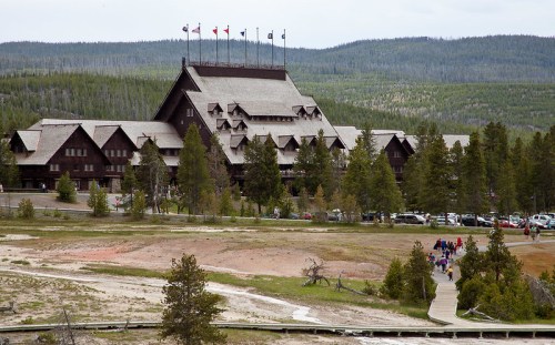 A large lodge with multiple flags, surrounded by trees and a walking path, set against a mountainous landscape.