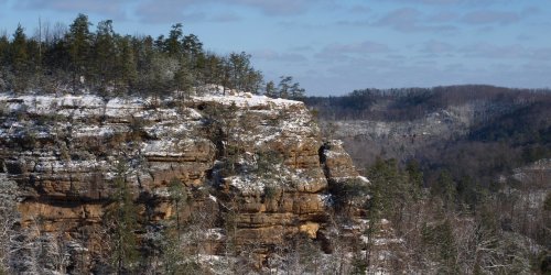 Snow-covered cliffs rise above a forested valley under a clear blue sky, showcasing a serene winter landscape.