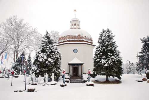 A snow-covered church with a dome, surrounded by evergreen trees and festive decorations in a winter landscape.