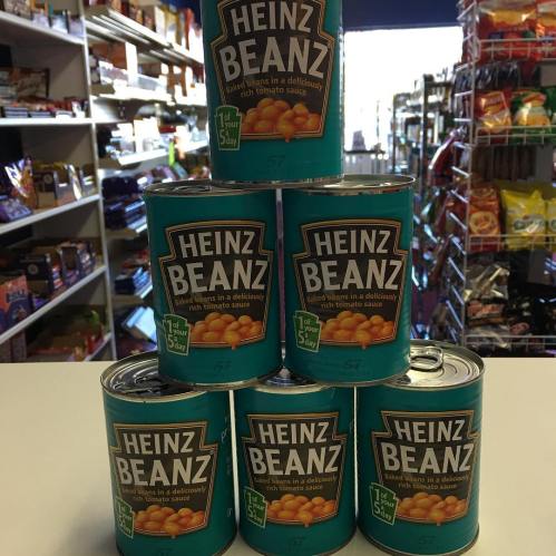 A stack of six cans of Heinz Baked Beans on a store counter, with shelves of snacks in the background.