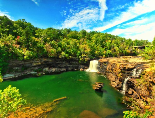 A scenic view of a waterfall cascading into a green pool, surrounded by lush trees and a blue sky with clouds.