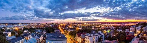 Panoramic view of a city skyline at sunset, with vibrant clouds and lights reflecting on the water.