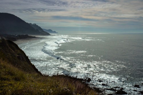 A serene coastal view with gentle waves, cliffs, and a cloudy sky reflecting sunlight over the ocean.