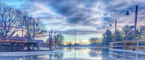 A serene ice rink surrounded by trees and lampposts, reflecting a cloudy sky at dusk.