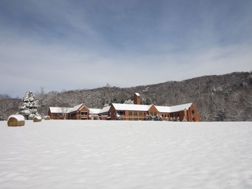 A snow-covered landscape featuring a large, rustic building surrounded by trees and hay bales under a blue sky.