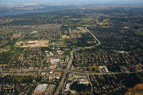 Aerial view of a suburban area with residential neighborhoods, commercial buildings, and green spaces under a clear sky.