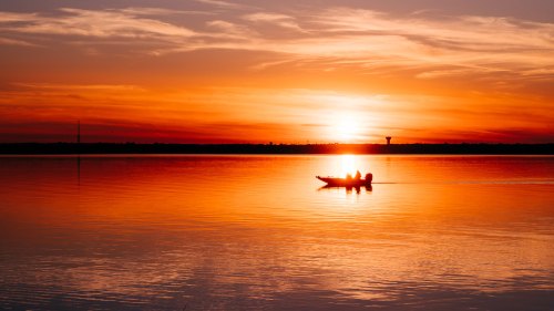 A silhouette of a boat on calm water at sunset, with vibrant orange and yellow hues in the sky.