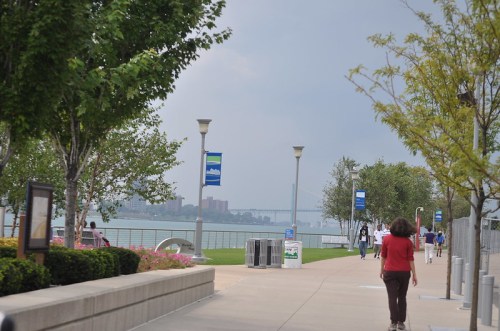 A scenic waterfront path lined with trees, benches, and lampposts, with people walking and a bridge in the background.