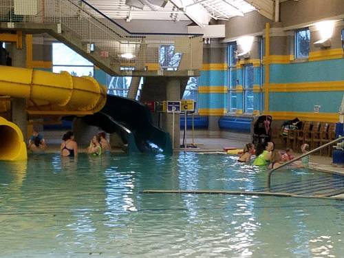 Indoor swimming pool with a yellow slide, people swimming, and lounging around in a vibrant, family-friendly environment.