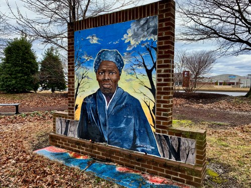 A mural of a historical figure, framed by brick, set in a park with trees and a cloudy sky in the background.