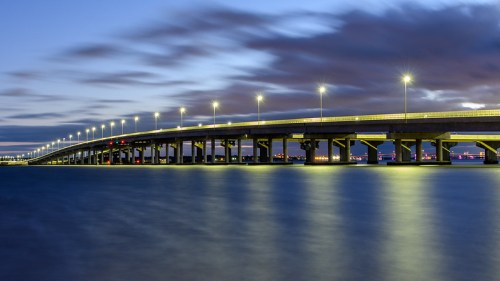 A long bridge illuminated by lights stretches over calm water at dusk, with clouds in the sky above.