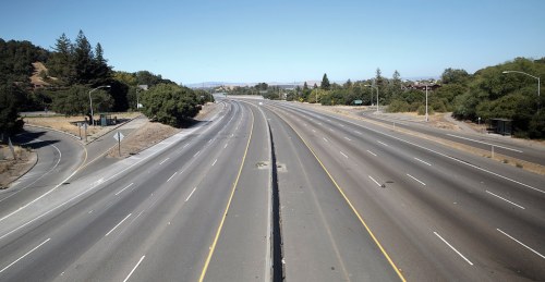 A wide, empty highway stretches into the distance, flanked by trees and clear blue skies.