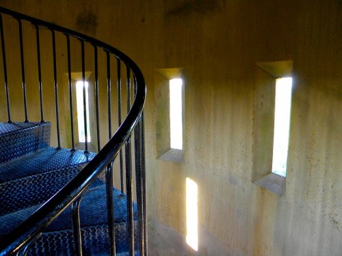 A spiral staircase with a metal railing, illuminated by narrow windows in a textured wall.
