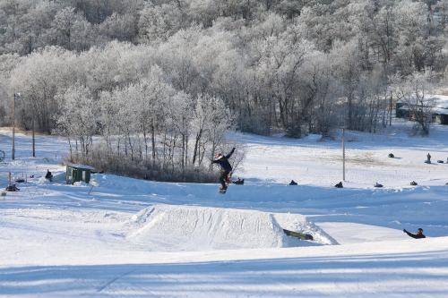 A snowboarder jumps over a snow ramp, surrounded by a snowy landscape and frosted trees in the background.