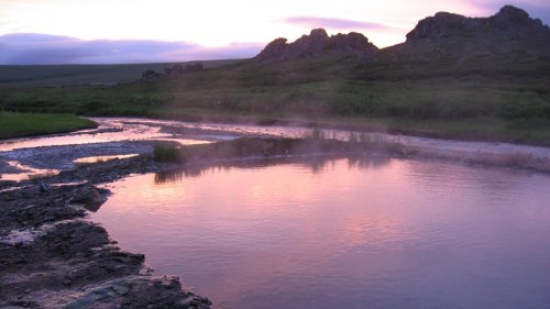 A serene landscape featuring a calm river reflecting a pink sunset, surrounded by rocky hills and lush greenery.