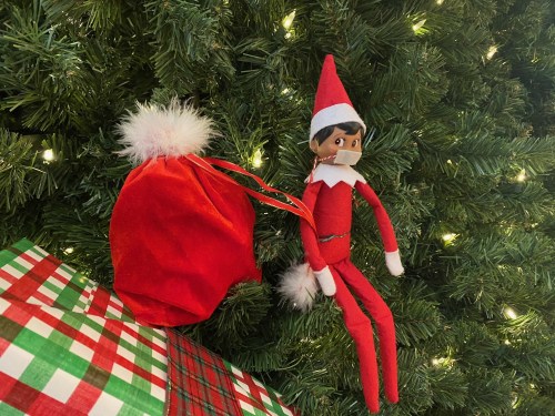 An elf doll with a red outfit and hat sits on a Christmas tree, next to a small red sack and wrapped gifts.