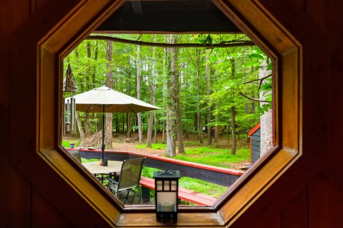 View through an octagonal window showcasing a lush green forest and outdoor seating area with an umbrella.