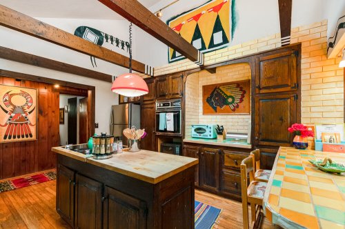 A cozy kitchen with wooden beams, colorful artwork, and a central island with a pink pendant light.