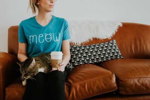 A person in a blue "meow" shirt sits on a couch with a cat and a coffee cup, surrounded by cat-themed decor.