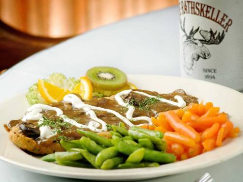 A plate of beef with gravy, served with carrots, green beans, kiwi, orange slices, and a drink in the background.