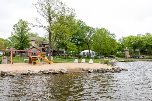 A sandy beach by a lake with a playground, chairs, and green trees in the background on a cloudy day.