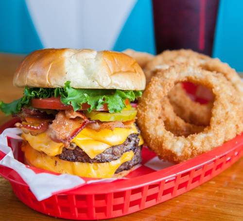 A stacked cheeseburger with bacon, lettuce, and tomato, served with onion rings in a red basket.