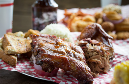 A platter of barbecue ribs, pulled pork, cornbread, coleslaw, and fries, served with a drink in the background.
